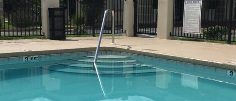 A swimming pool with a metal ladder, surrounded by a fence and a building with balconies.