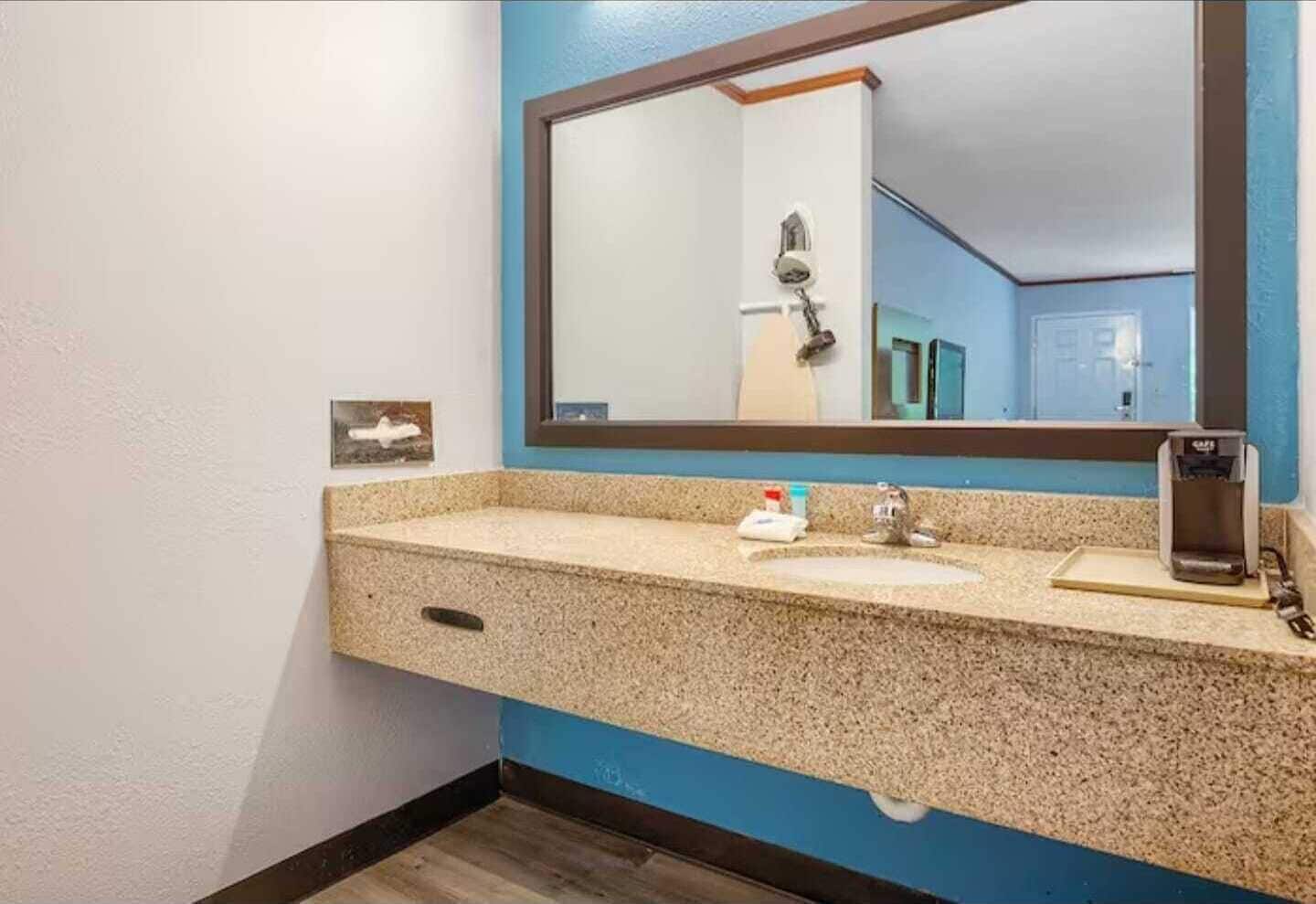 A bathroom with a granite countertop, a sink, a mirror, a towel dispenser, and a coffee maker.