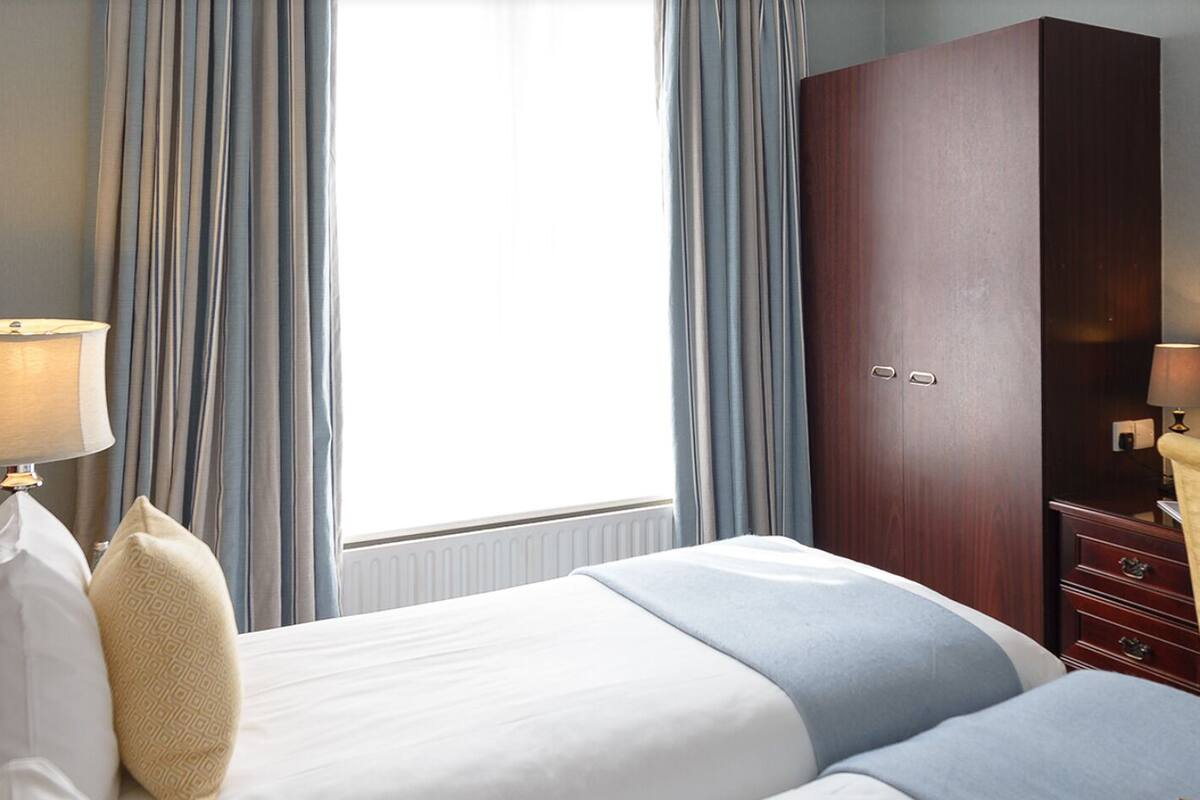 Ein Hotelzimmer mit Bett, Nachttisch, Lampe, Kleiderschrank und einem Fenster mit Vorhängen.