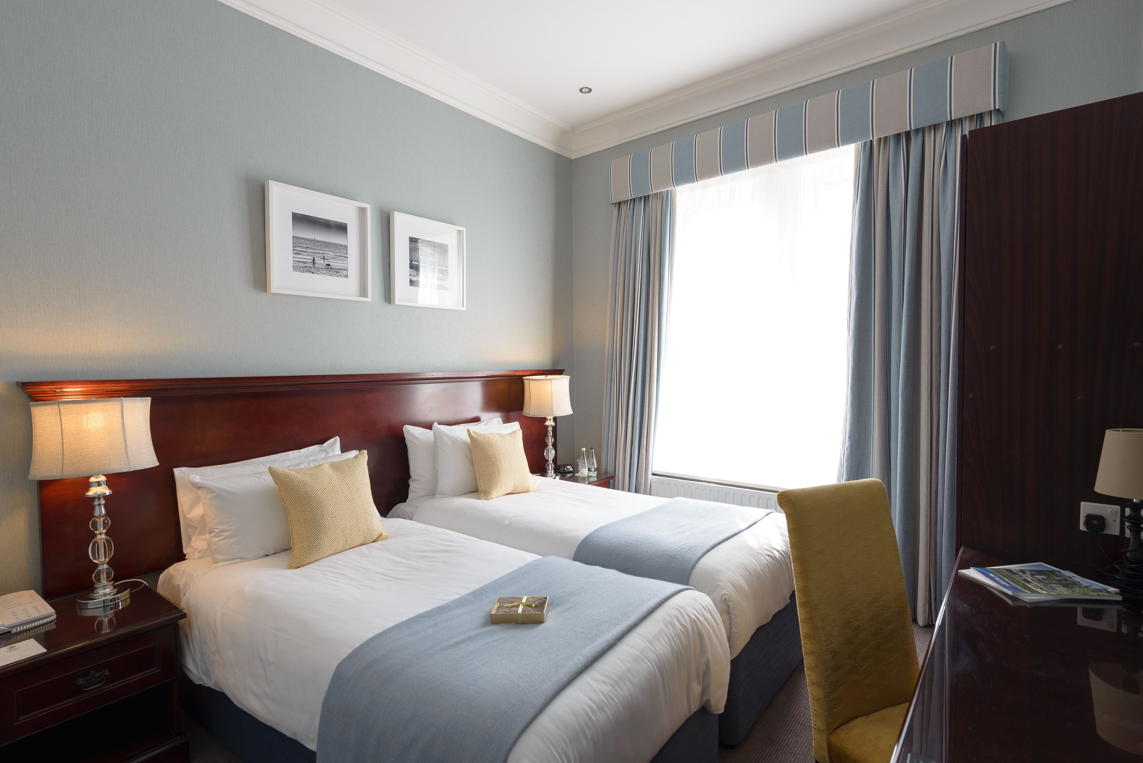 Ein Hotelzimmer mit zwei Betten, einem Holzkopfende, einem Nachttisch mit Lampe, einem Schreibtisch mit Stuhl und einem großen Fenster mit Vorhängen.