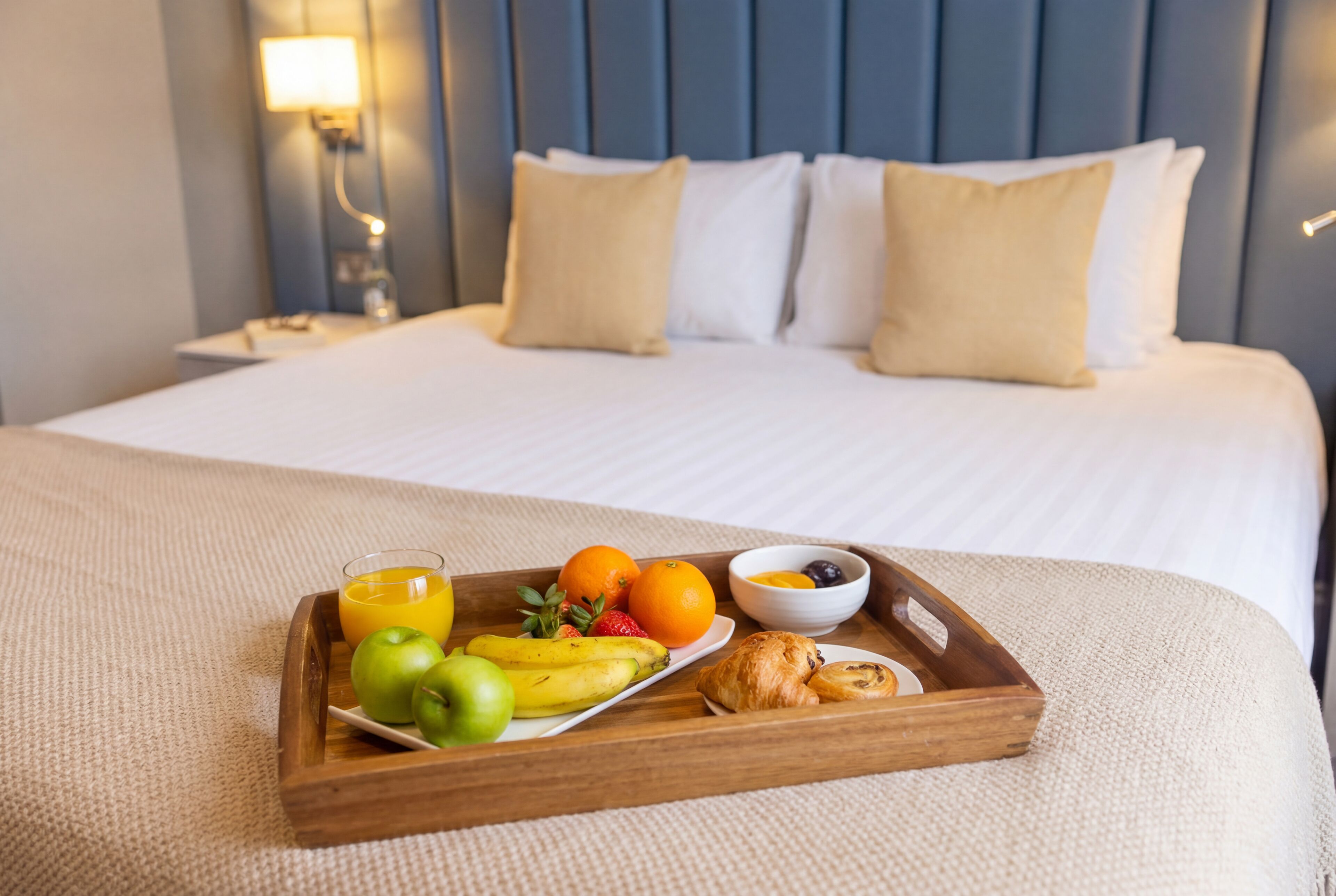 A neatly made bed with a tray of breakfast items including fruits, a croissant, and a glass of orange juice.