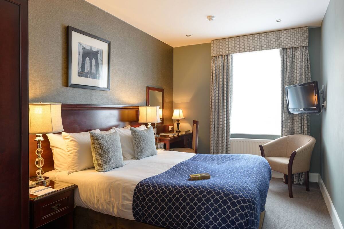 Ein Hotelzimmer mit einem Bett, Nachttischlampen, einem Schreibtisch mit Lampe, einem Sessel, einem Fernseher und einem Fenster mit Vorhängen.