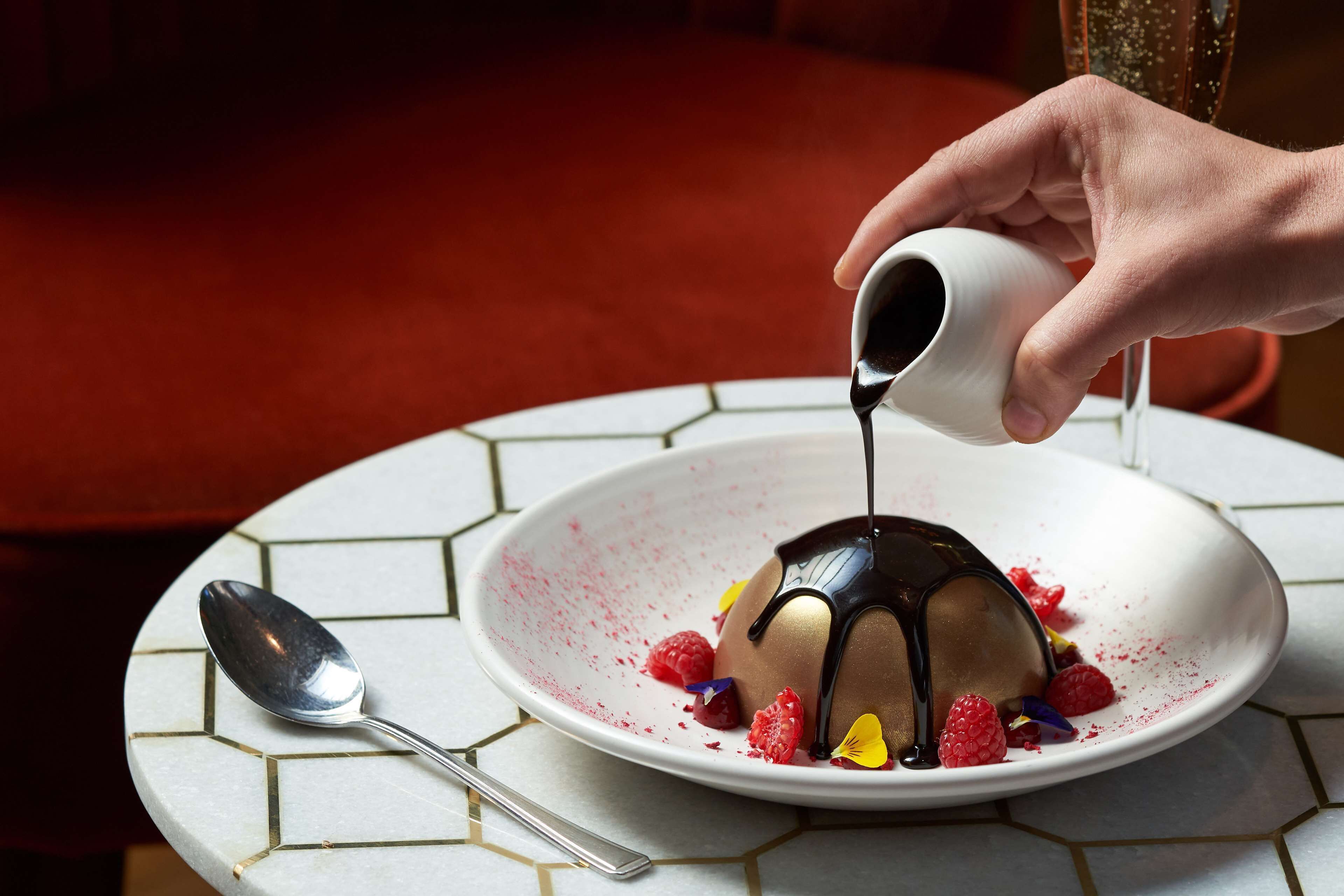 A dessert on a white plate with a spoon and a cup pouring chocolate sauce.