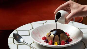 A dessert on a white plate with a spoon and a cup pouring chocolate sauce.
