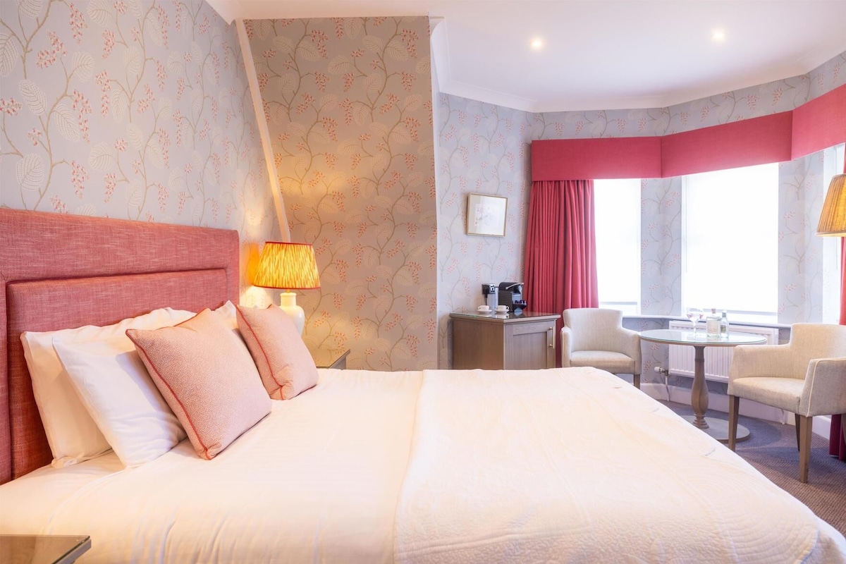A hotel room with a large bed, a desk with a chair, a small table, and a window with curtains.