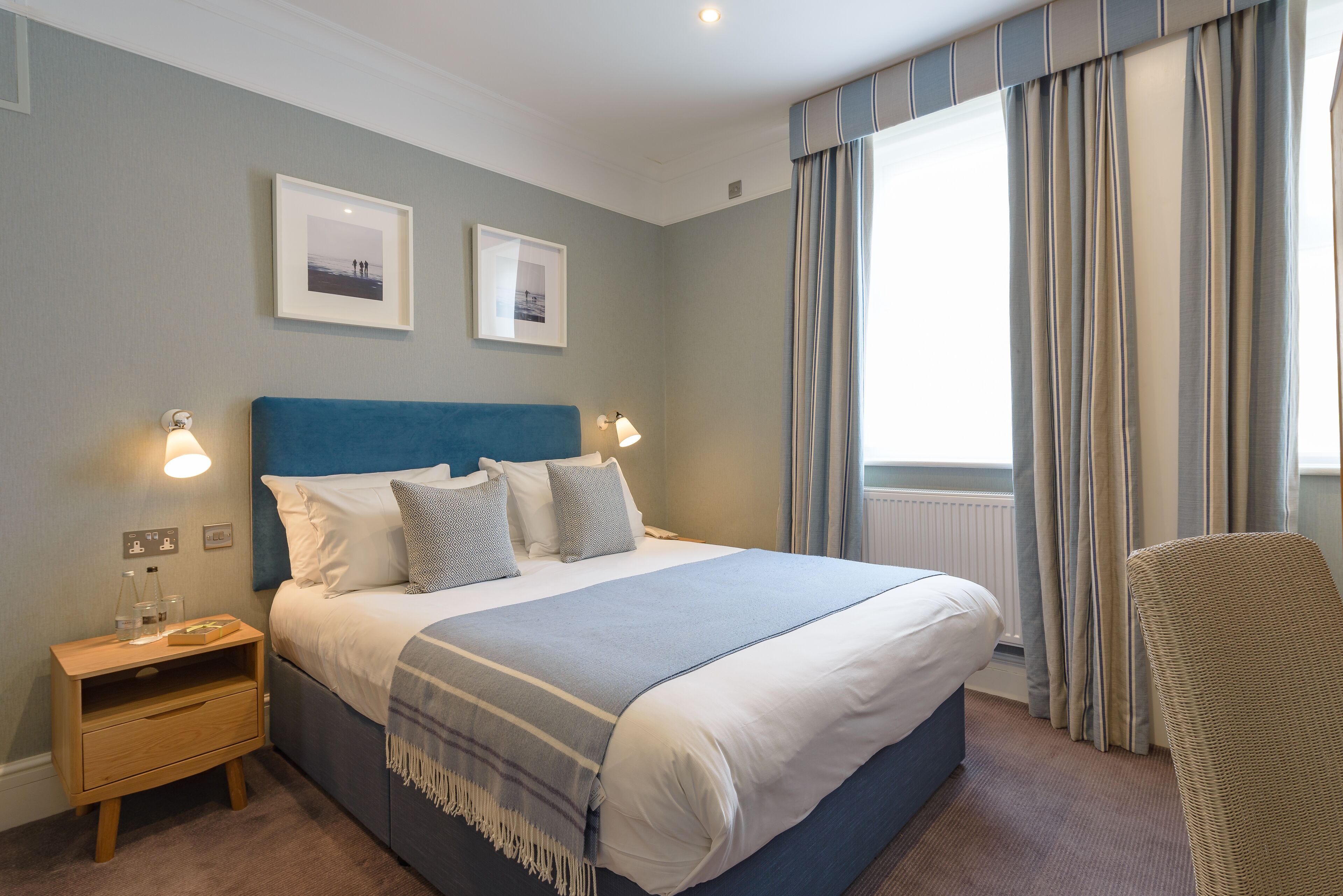 A neatly made bed with a blue headboard, a bedside table with a lamp, and two framed pictures on the wall.