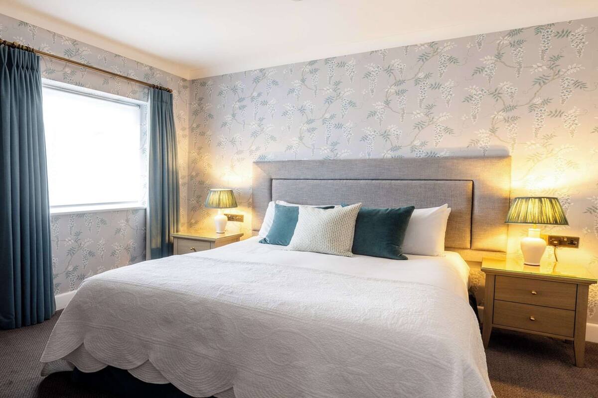 Ein ordentlich bezogenes Bett mit weißer Bettdecke und zwei Kissen, begleitet von Nachttischen mit Lampen, in einem Zimmer mit floralem Wandpapier und einem großen Fenster mit blauen Vorhängen.