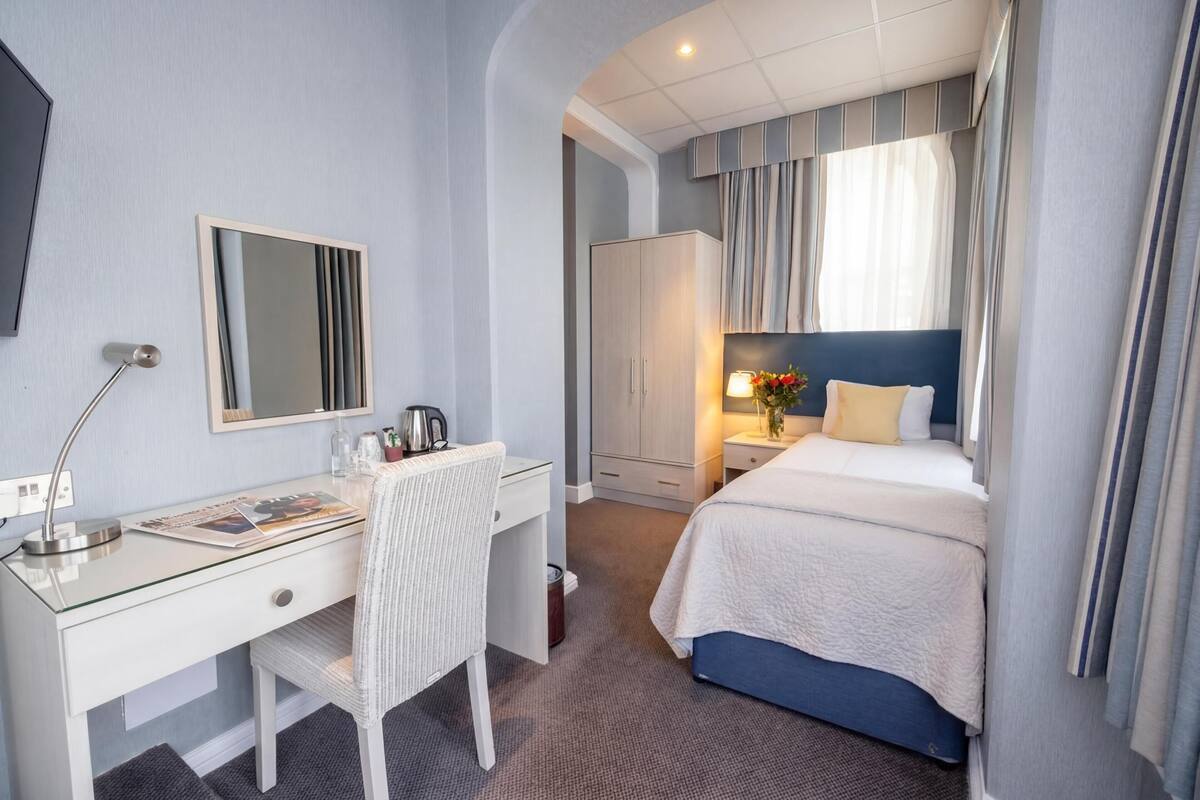 Ein Hotelzimmer mit einem Bett, einem Schreibtisch mit Stuhl, einem Kleiderschrank, einem Spiegel und einem Nachttisch mit Lampe und Blumen.