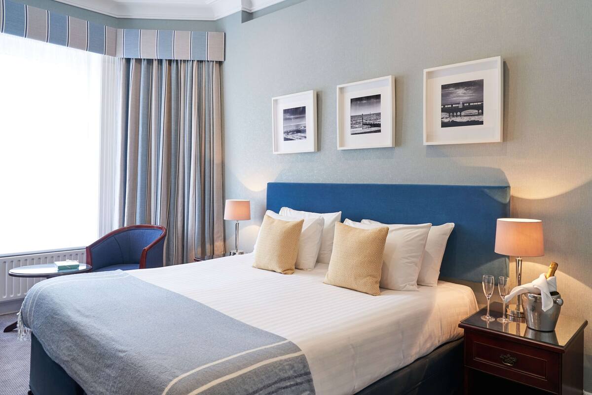 Ein Hotelzimmer mit einem großen Bett, zwei Nachttischlampen, einem Nachttisch mit einer Champagnerflasche und Gläsern, einem Sessel und drei gerahmten Bildern an der Wand.