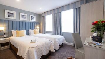 A hotel room with two beds, a desk, and a chair. There are framed pictures on the wall, a vase with flowers, and a pair of shoes on the floor.