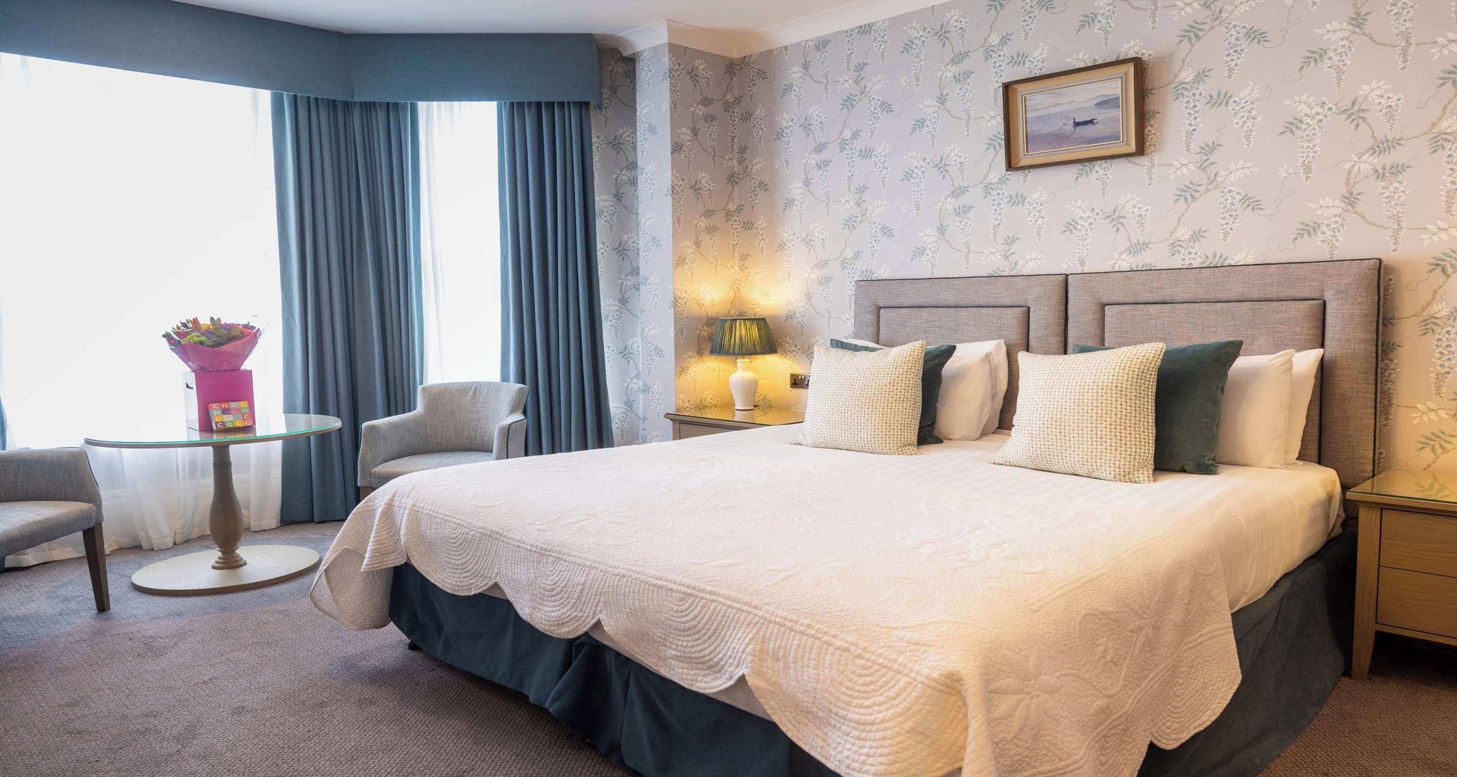 Een hotelkamer met een groot bed, een ronde glazen tafel, een stoel, een nachtkastje met een lamp en een raam met gordijnen.