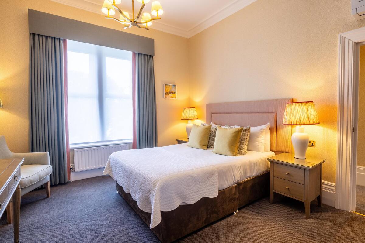 Ein Hotelzimmer mit Bett, Nachttischlampen, einem Sessel und einem Fenster mit Vorhängen.