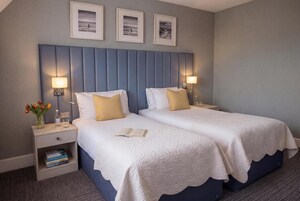 Habitación de hotel con dos camas, una mesita de noche con lámpara, un florero con flores y cuadros en la pared.