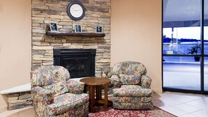 A cozy room with a stone fireplace, two floral armchairs, a small wooden table, and a large window offering a view of the outside. - Ramada by Wyndham Watertown/Thousand Islands NY (Watertown)