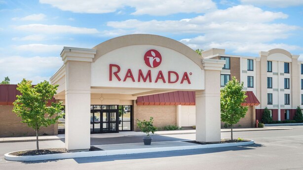 A Ramada hotel entrance with a clear sky, a red awning, and two potted plants. - Ramada by Wyndham Watertown/Thousand Islands NY (Watertown)