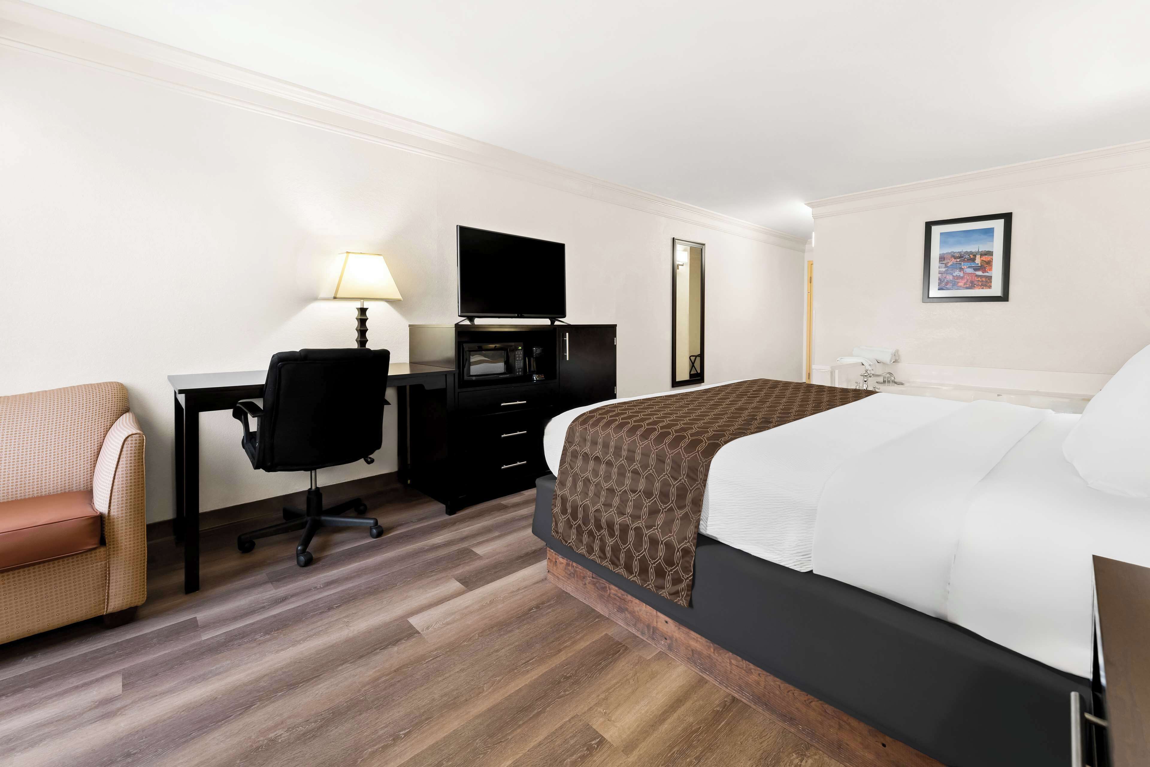 A hotel room with a bed, a desk with a computer, a chair, a lamp, a flat-screen TV, and a framed picture on the wall.