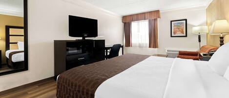 A hotel room with a large bed, a flat-screen TV mounted on the wall, a desk with a chair, a window with curtains, and a lamp on the side table.