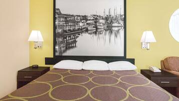 A hotel room with a bed, two bedside tables, a nightstand, a lamp, a chair, and a large framed picture of a harbor.