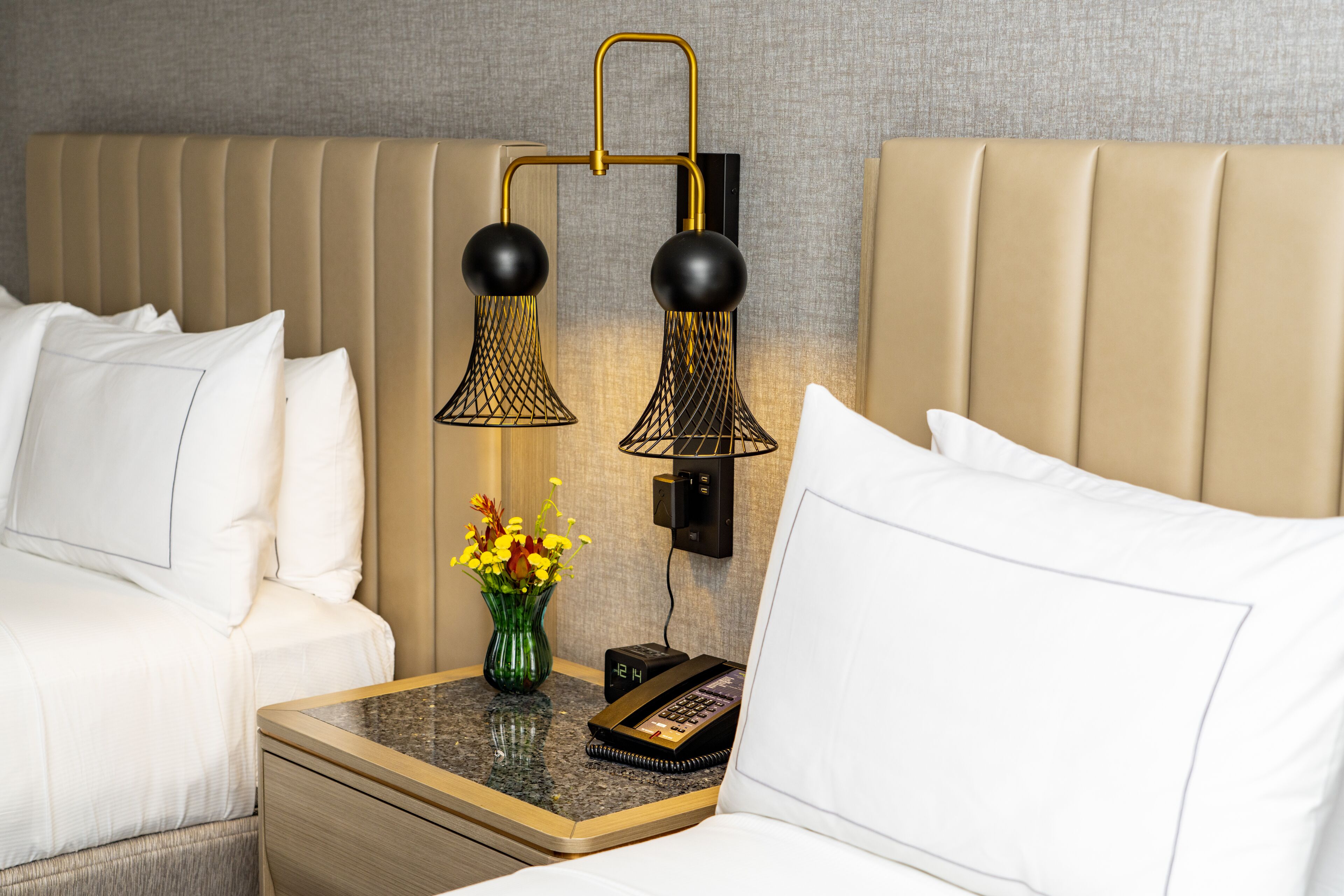 A hotel room with a bed, a nightstand with a phone, a vase with flowers, and a wall-mounted lamp.