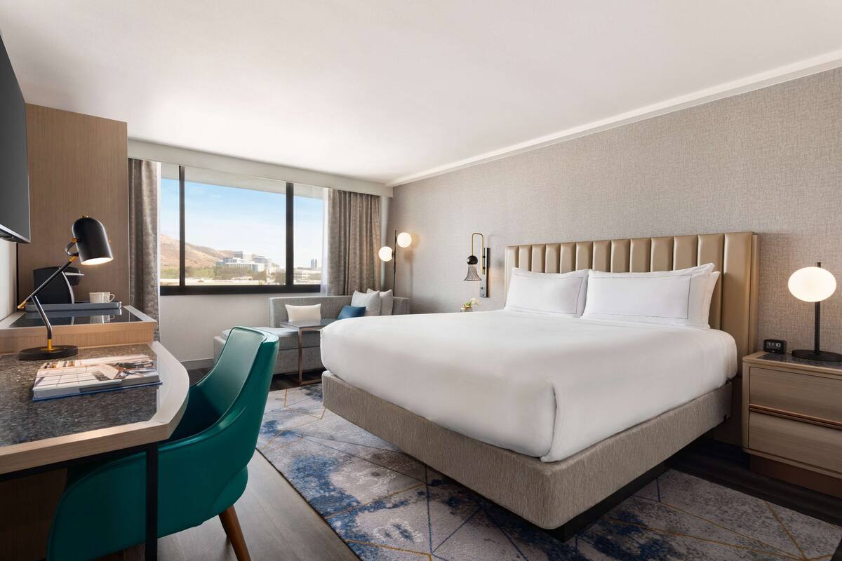 A hotel room with a large bed, a desk with a lamp, a chair, a nightstand, and a window with a view of the city.