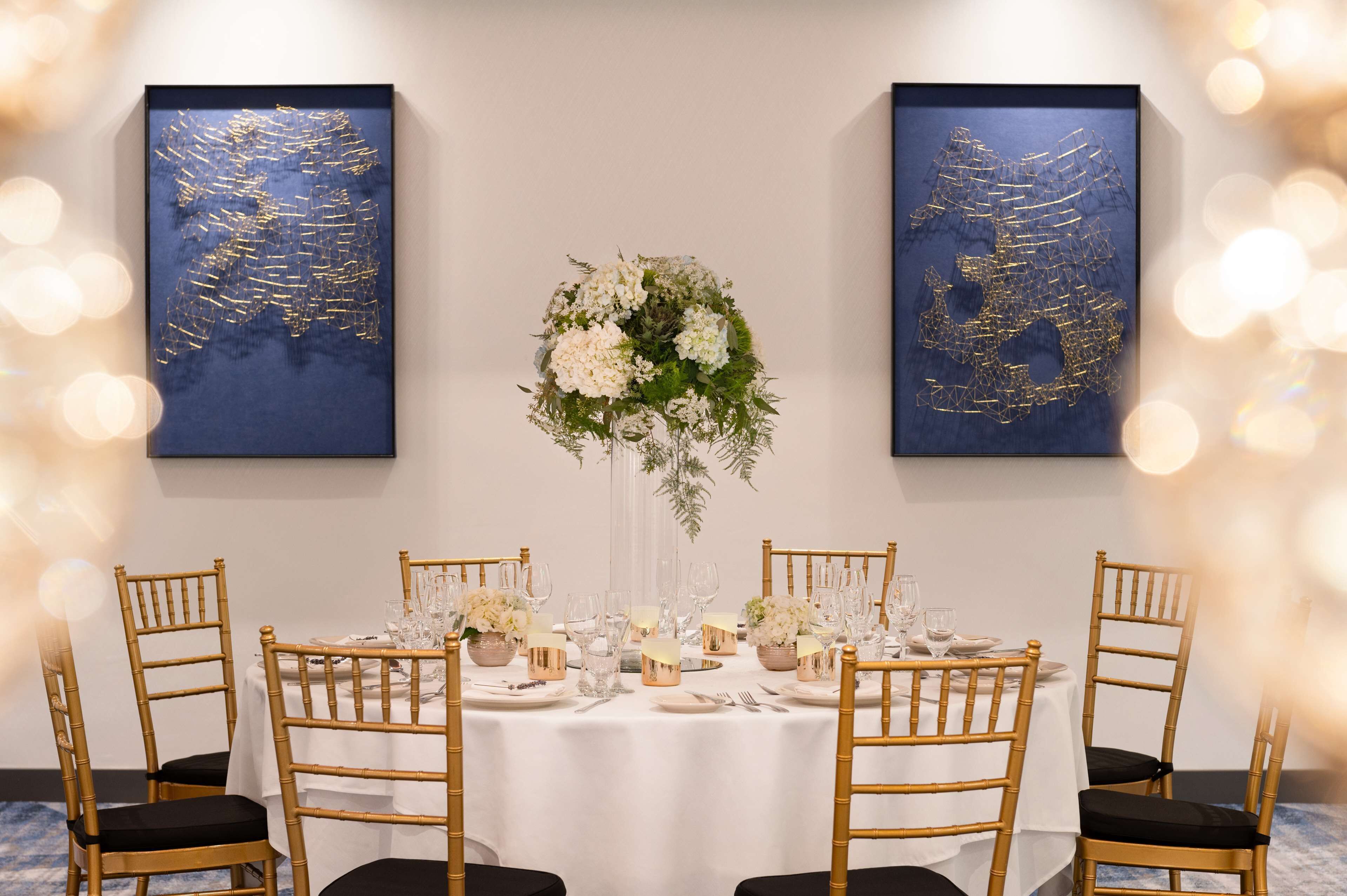 Mesa de comedor lista para seis personas, con sillas doradas, mantel blanco y centros de mesa florales.