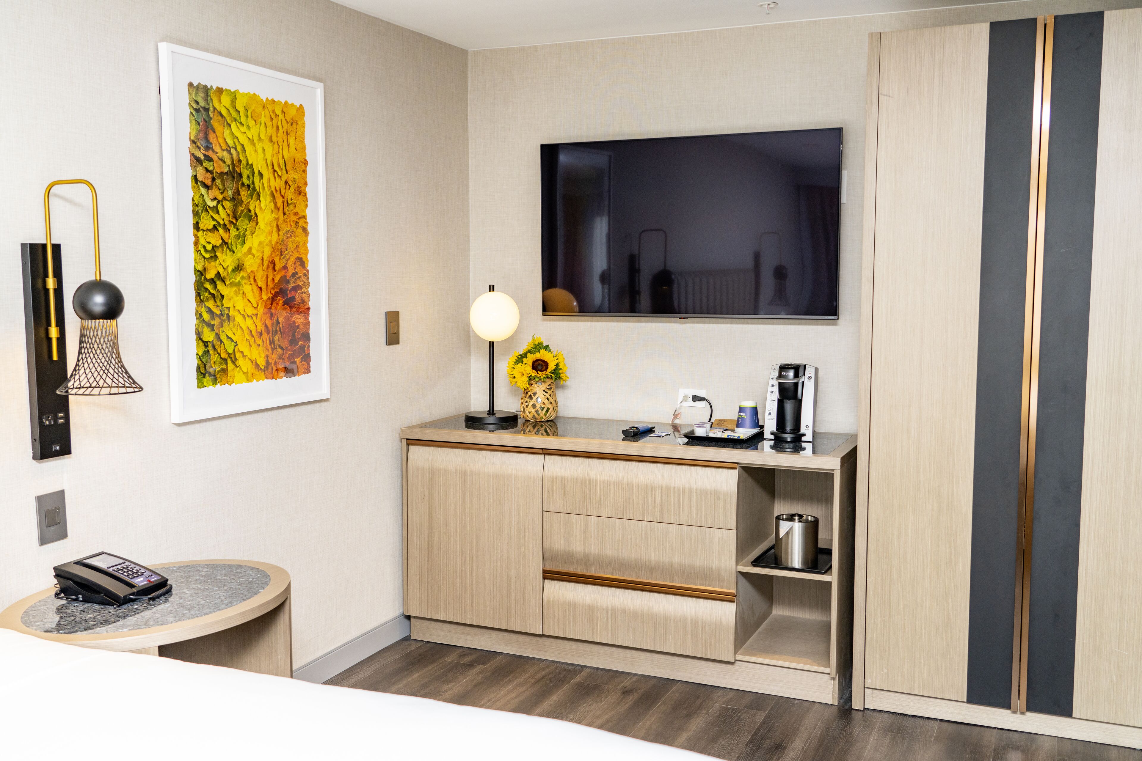 Une chambre d’hôtel moderne avec un meuble en bois, une table ronde avec un téléphone, une œuvre d’art encadrée, un vase avec un tournesol, un téléviseur à écran plat et une machine à café.