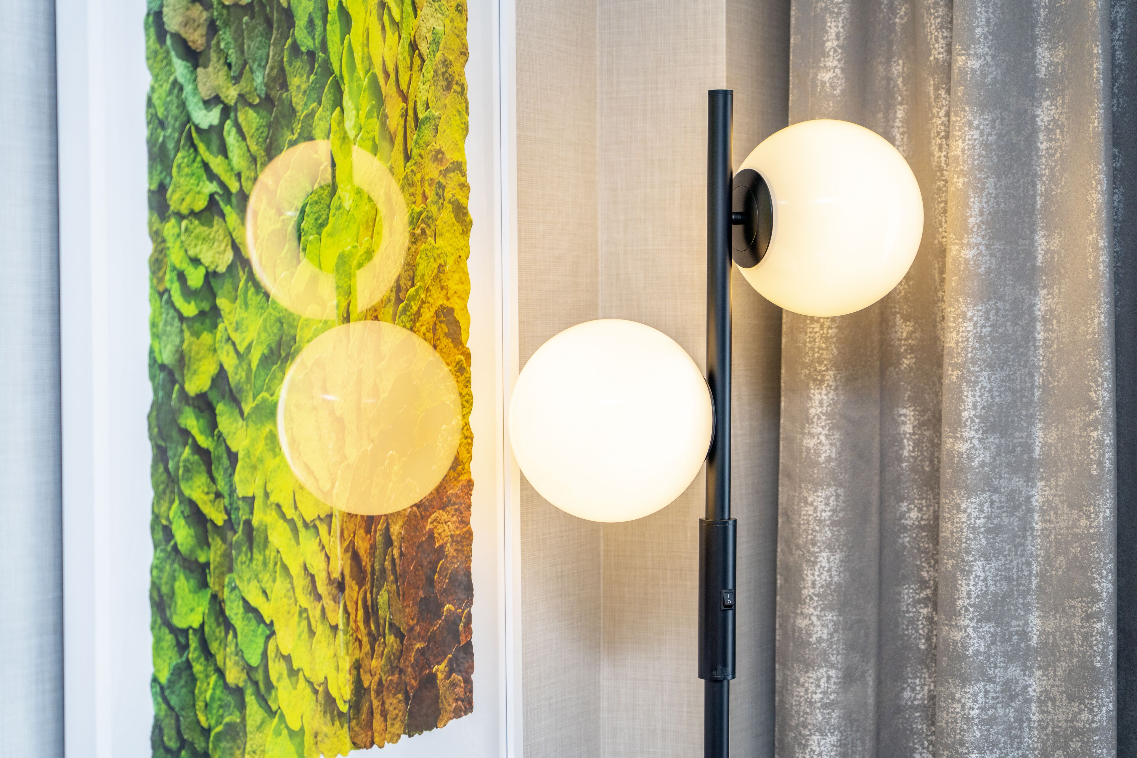 En vägg med en grön och gul målad väggmålning som har en tydlig struktur, en svart golvlampa med två sfäriska lampor och en grå gardin.