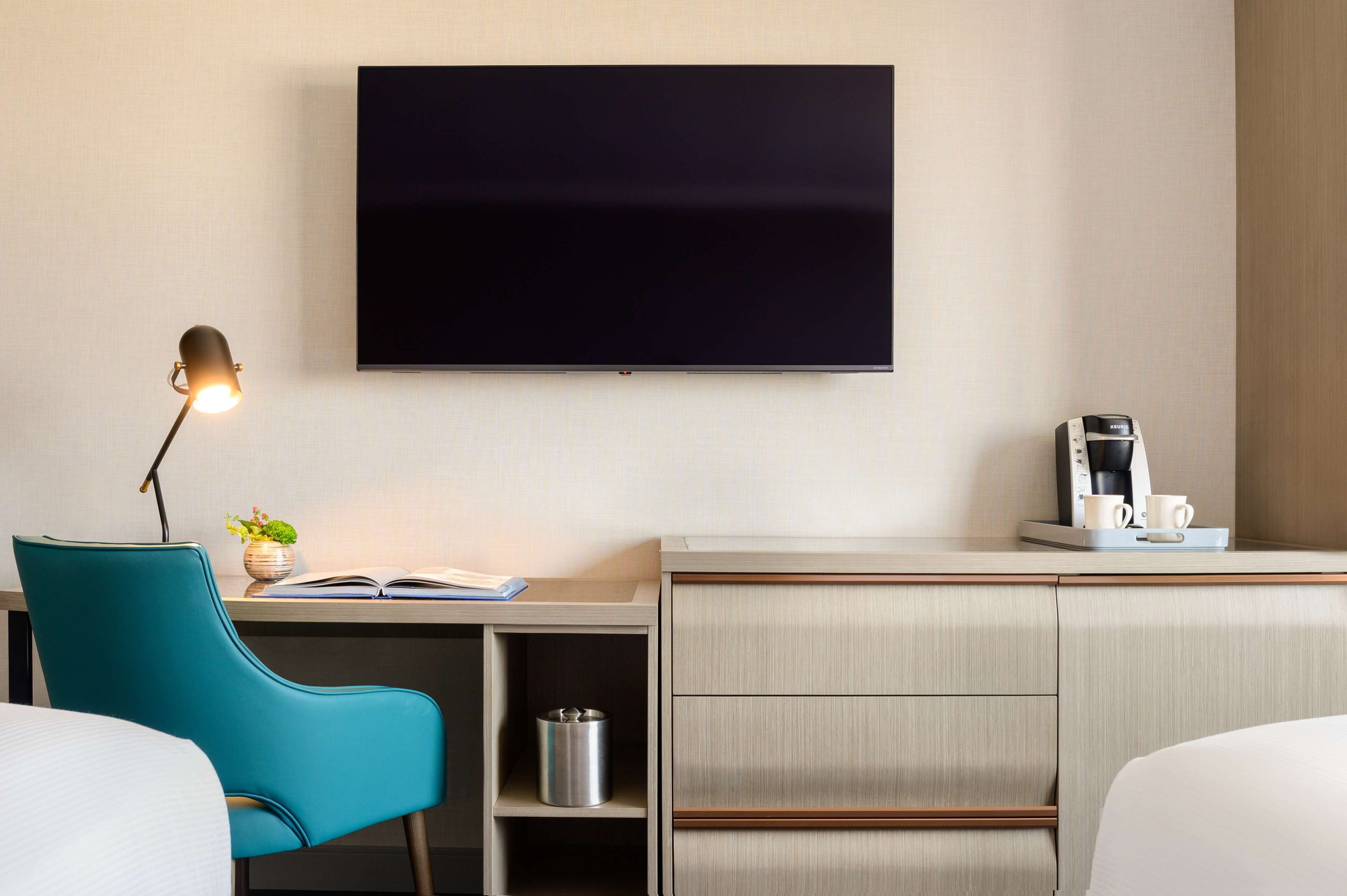 A hotel room with a flat-screen TV mounted on the wall, a desk with a lamp, a chair, a small plant, a coffee maker, and a bedside table with a cup and saucer.