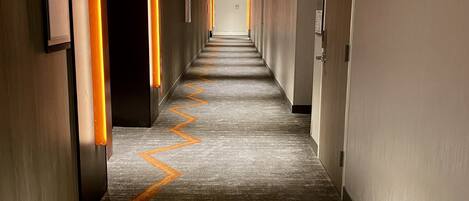 Un largo y estrecho pasillo de hotel con una alfombra estampada y lámparas fijadas a la pared.