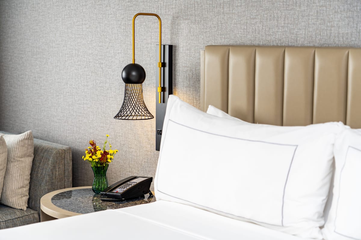 A neatly arranged hotel room with a bed, a bedside table with a lamp, a vase of flowers, and a telephone.