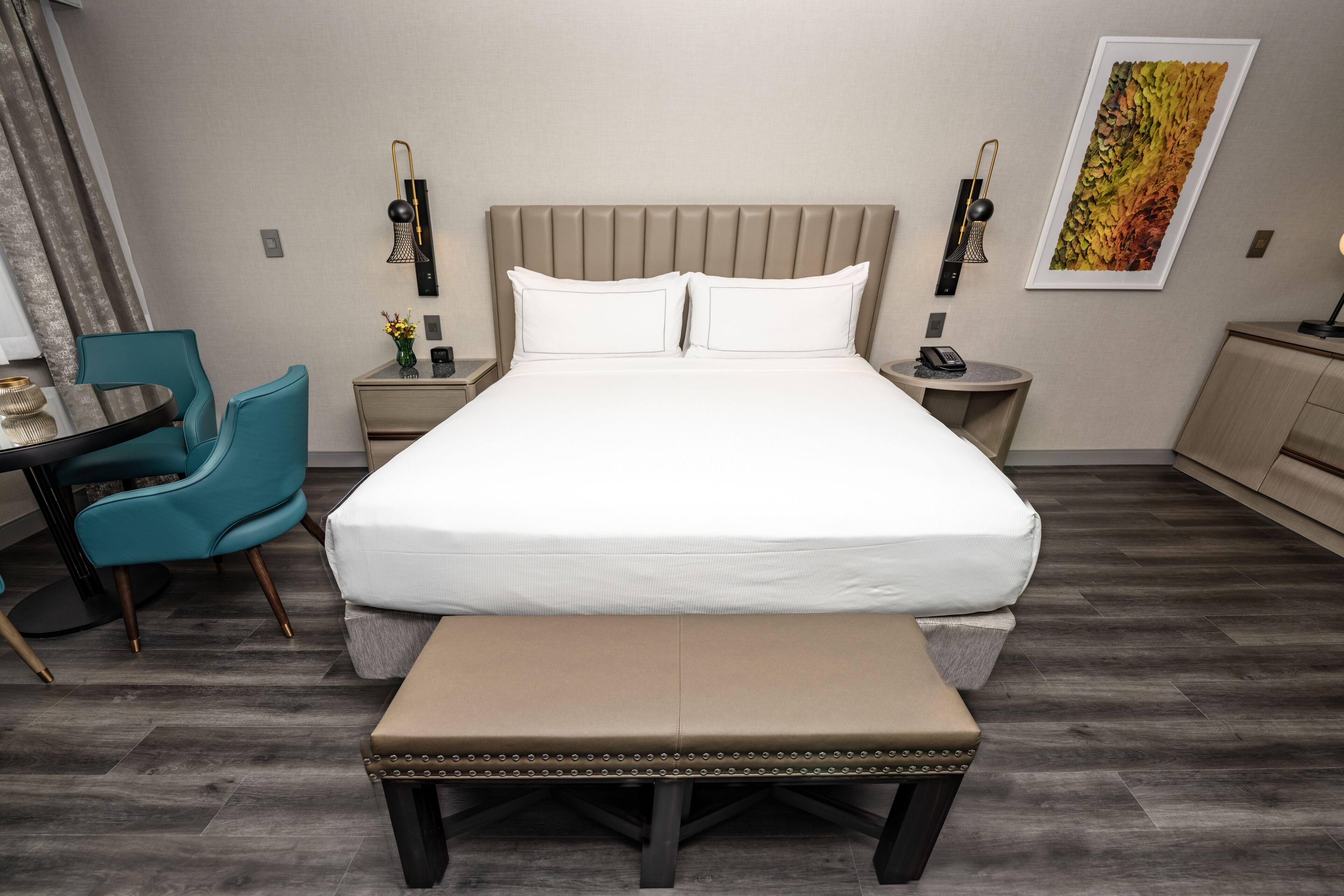 A hotel room with a large bed, a bench, a desk with a chair, a nightstand, and a wall-mounted lamp.