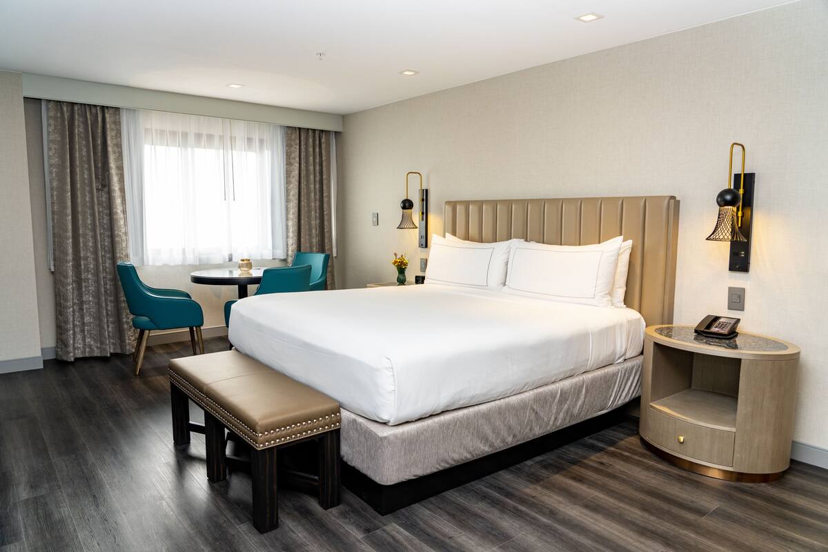 A hotel room with a large bed, a bedside table with a lamp, a small desk with a chair, and a bench.