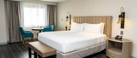 A hotel room with a large bed, a bedside table with a lamp, a small desk with a chair, and a bench.
