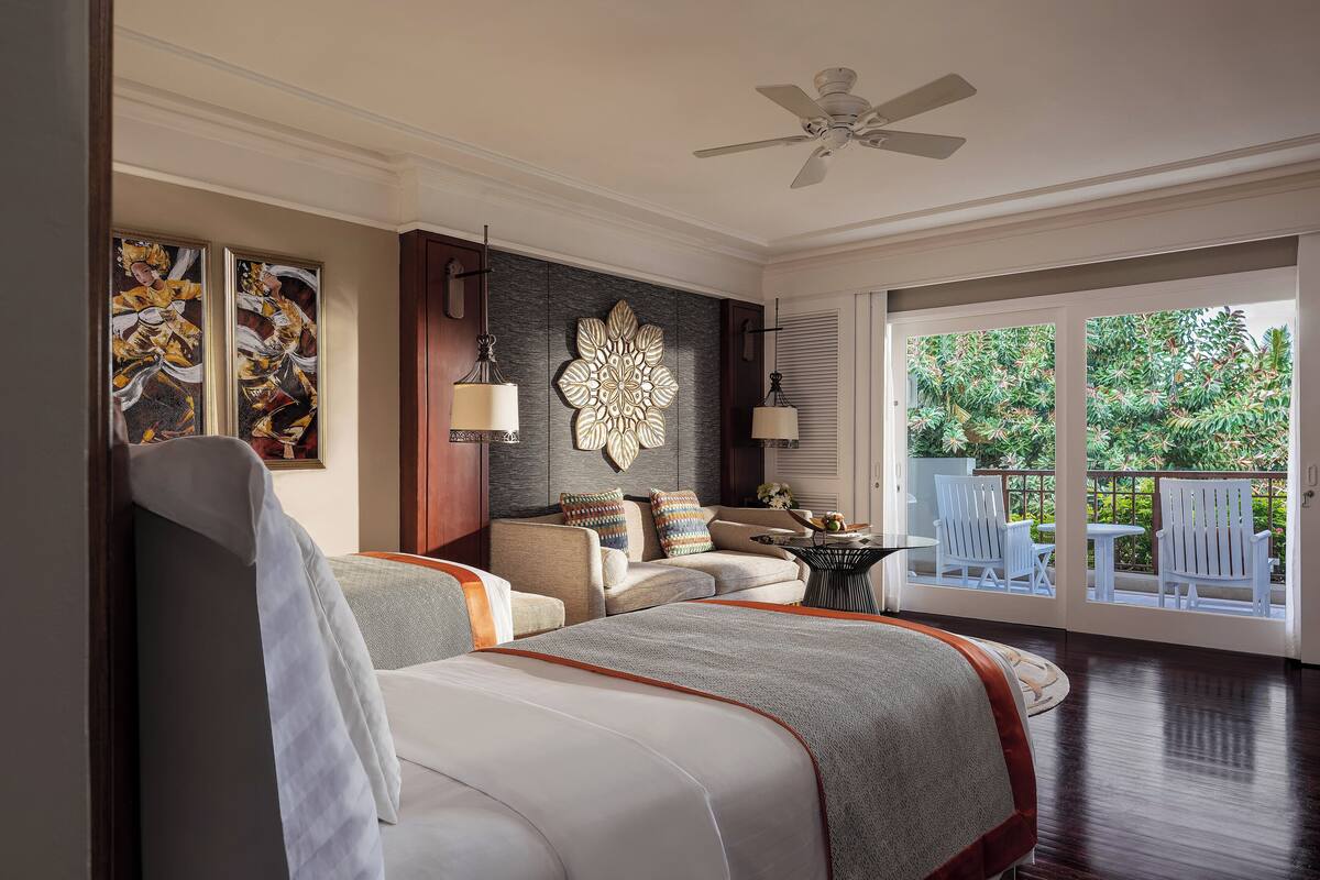A spacious bedroom with a large bed, a seating area with a sofa and chairs, a decorative wall piece, and a balcony with white chairs.