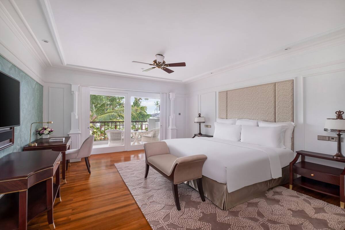 A hotel room with a large bed, a desk, a chair, a television, and a balcony with outdoor furniture.