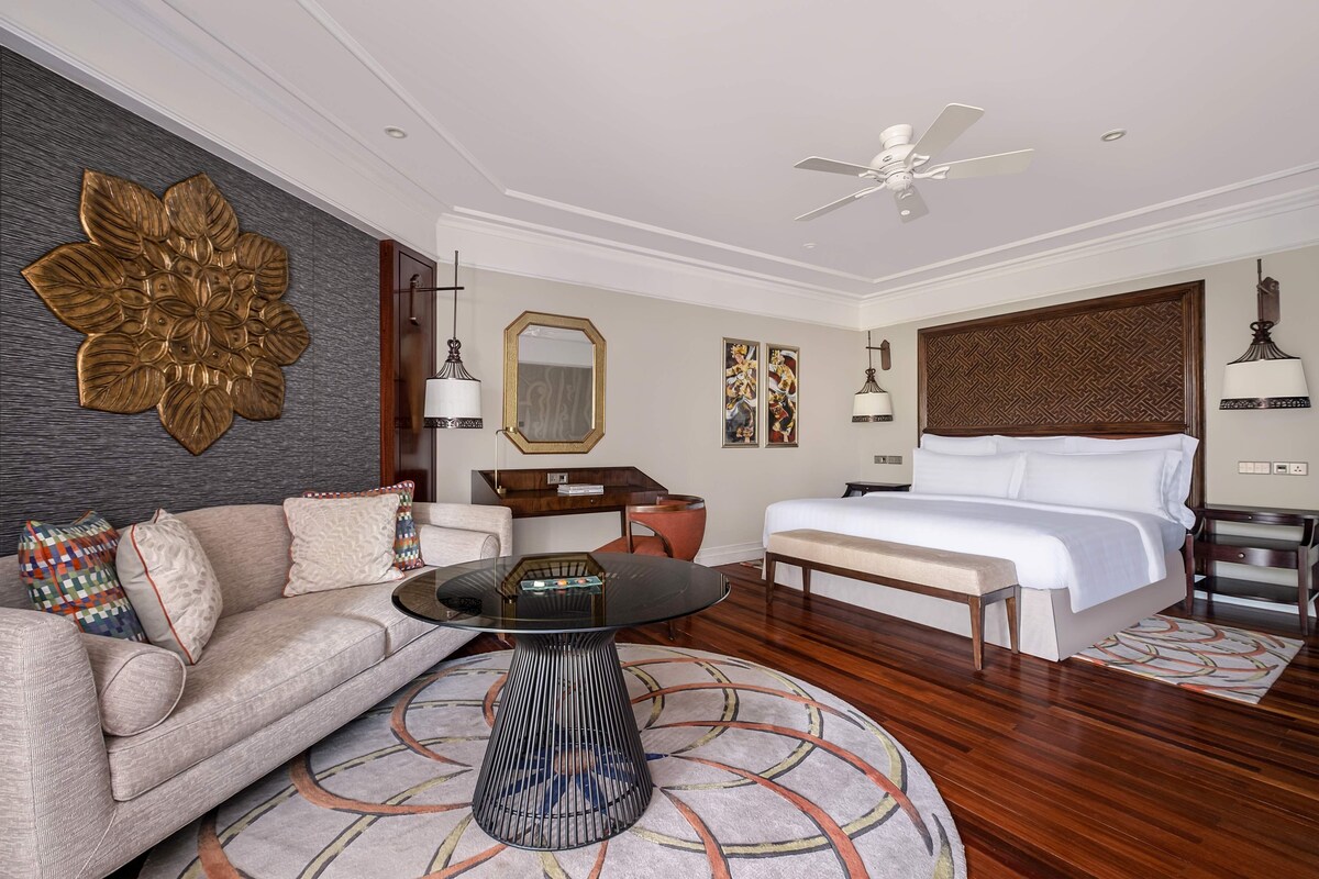 A hotel room with a large bed, a sofa, a coffee table, a ceiling fan, and a decorative wall piece.