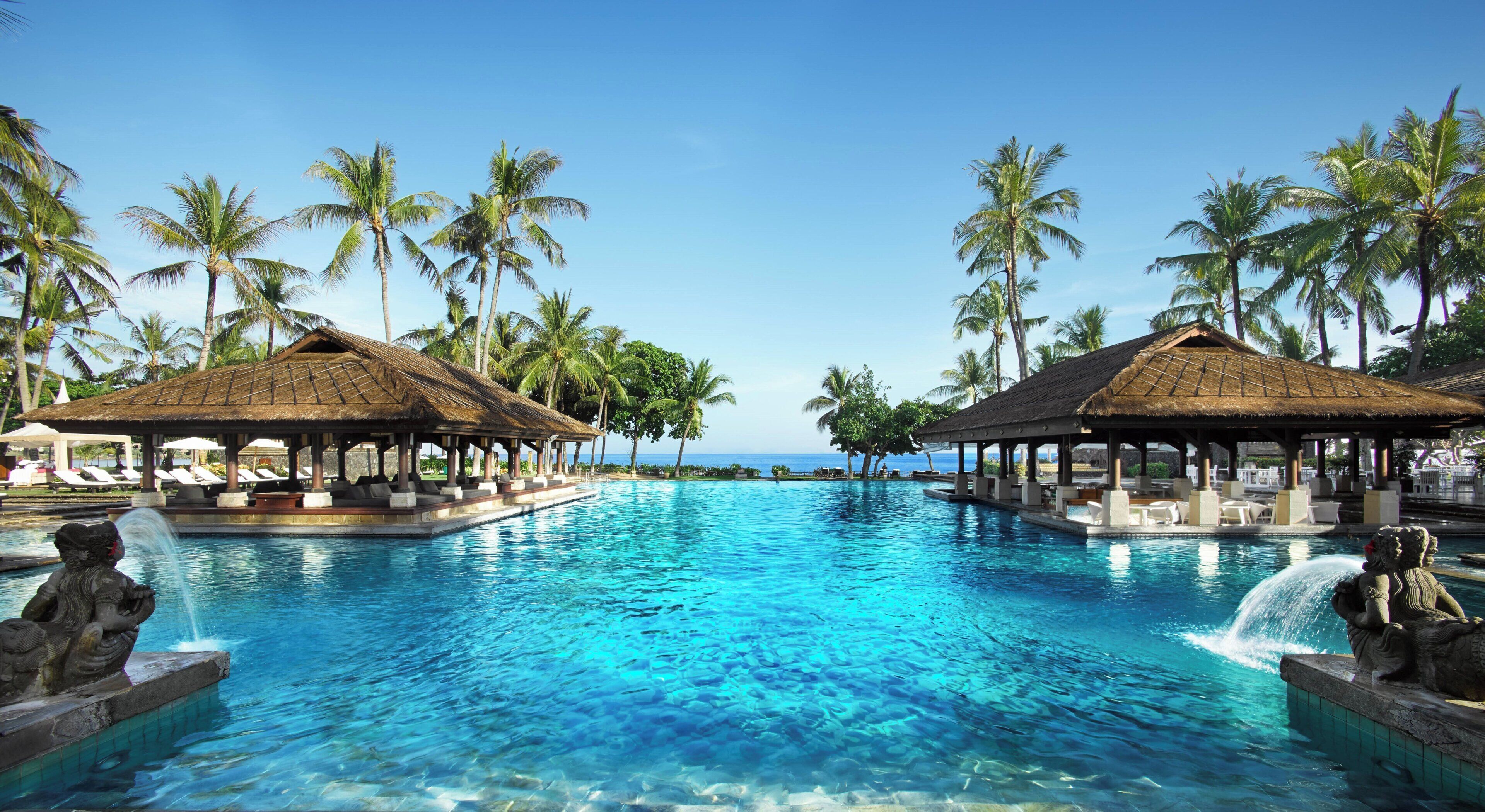 Una piscina de resort con agua azul cristalina, rodeada de palmeras y quioscos con techo de paja.