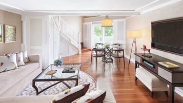 A spacious living room with a sofa, coffee table, and TV stand. There is a staircase leading to another level, and a dining area with chairs and a table.