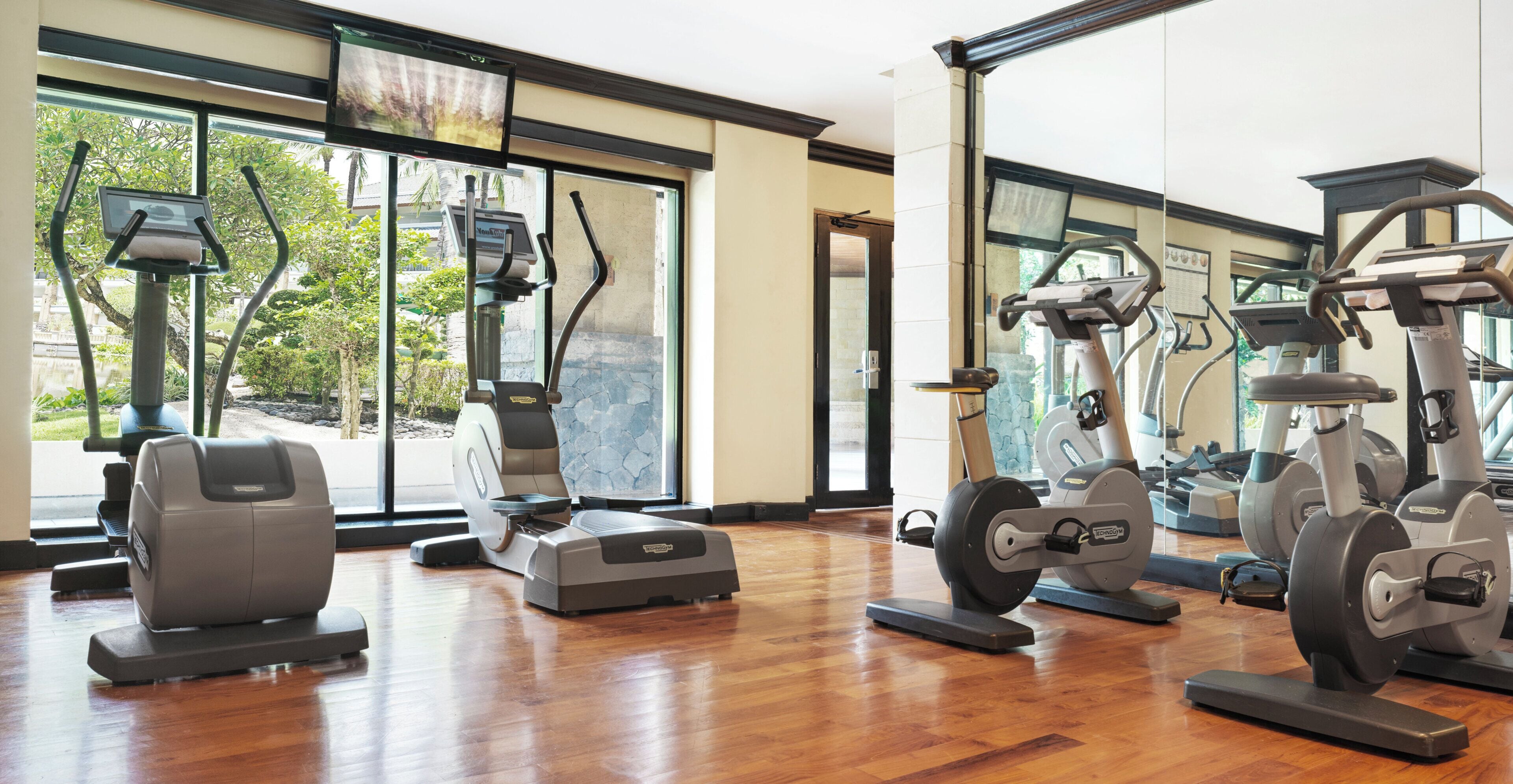 A gym with various exercise machines, large windows, and a wooden floor.