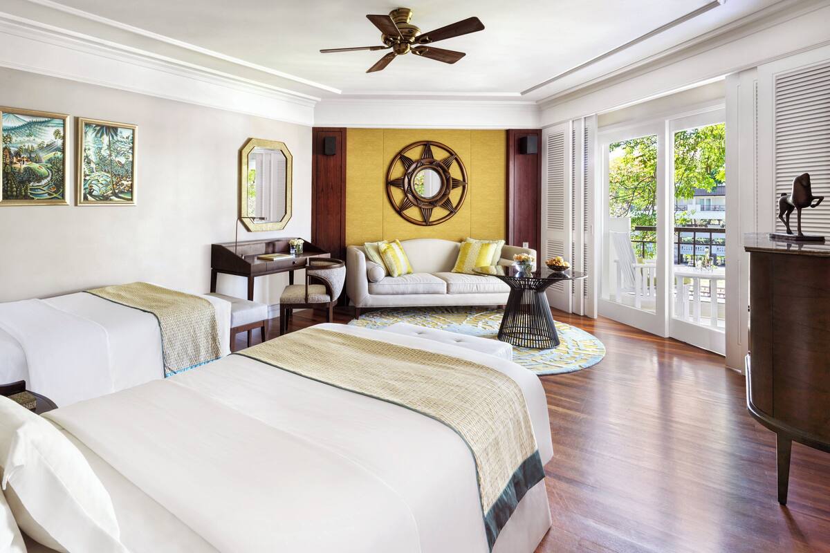 A hotel room with two beds, a desk, a sofa, a round coffee table, and a large window with a view of greenery.
