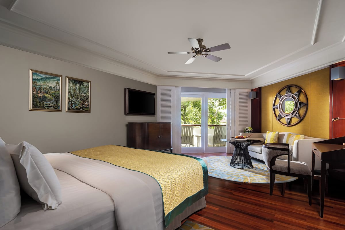 A hotel room with a bed, a desk, a chair, a ceiling fan, and a balcony with a view of greenery.