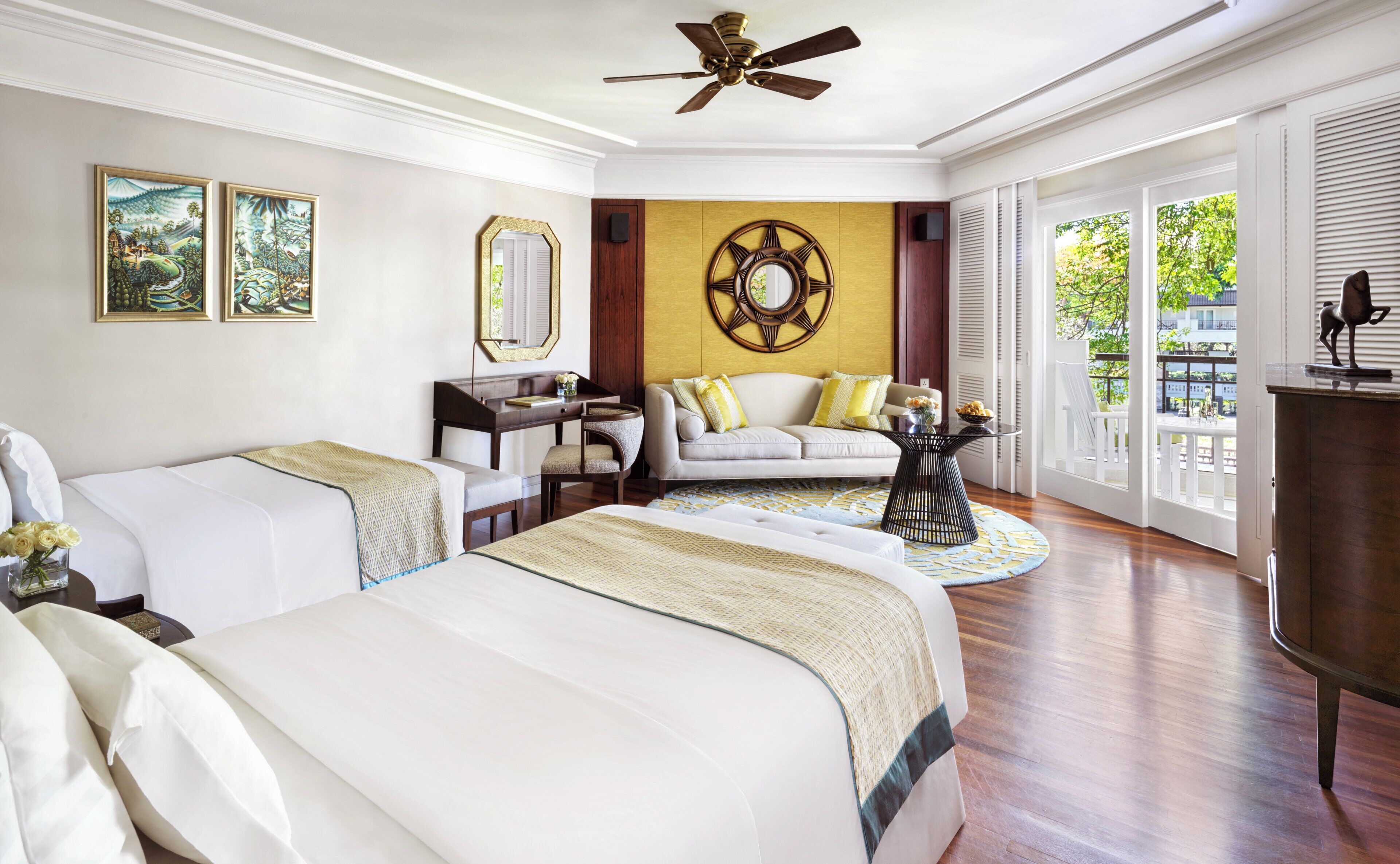 A hotel room with two beds, a desk, a sofa, a round coffee table, and a large window with a view of greenery.