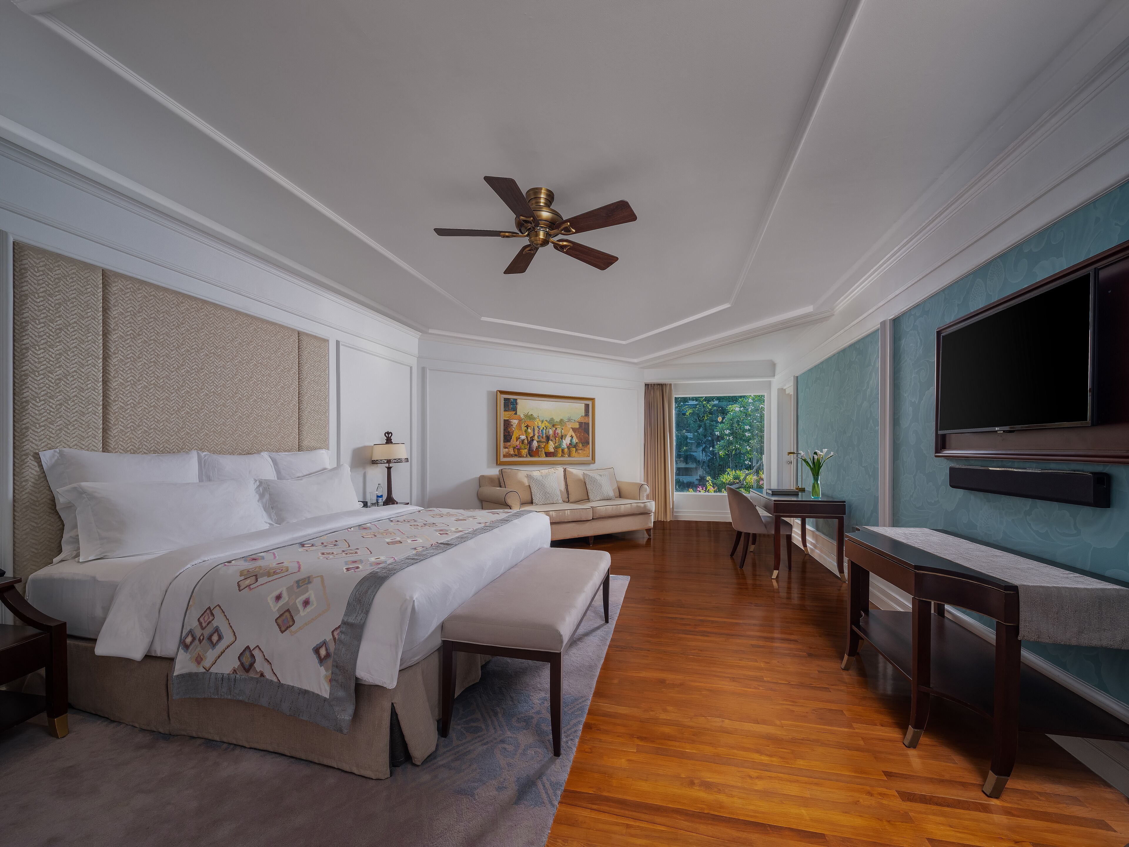 A spacious bedroom with a large bed, a ceiling fan, a TV, and a view of greenery outside.