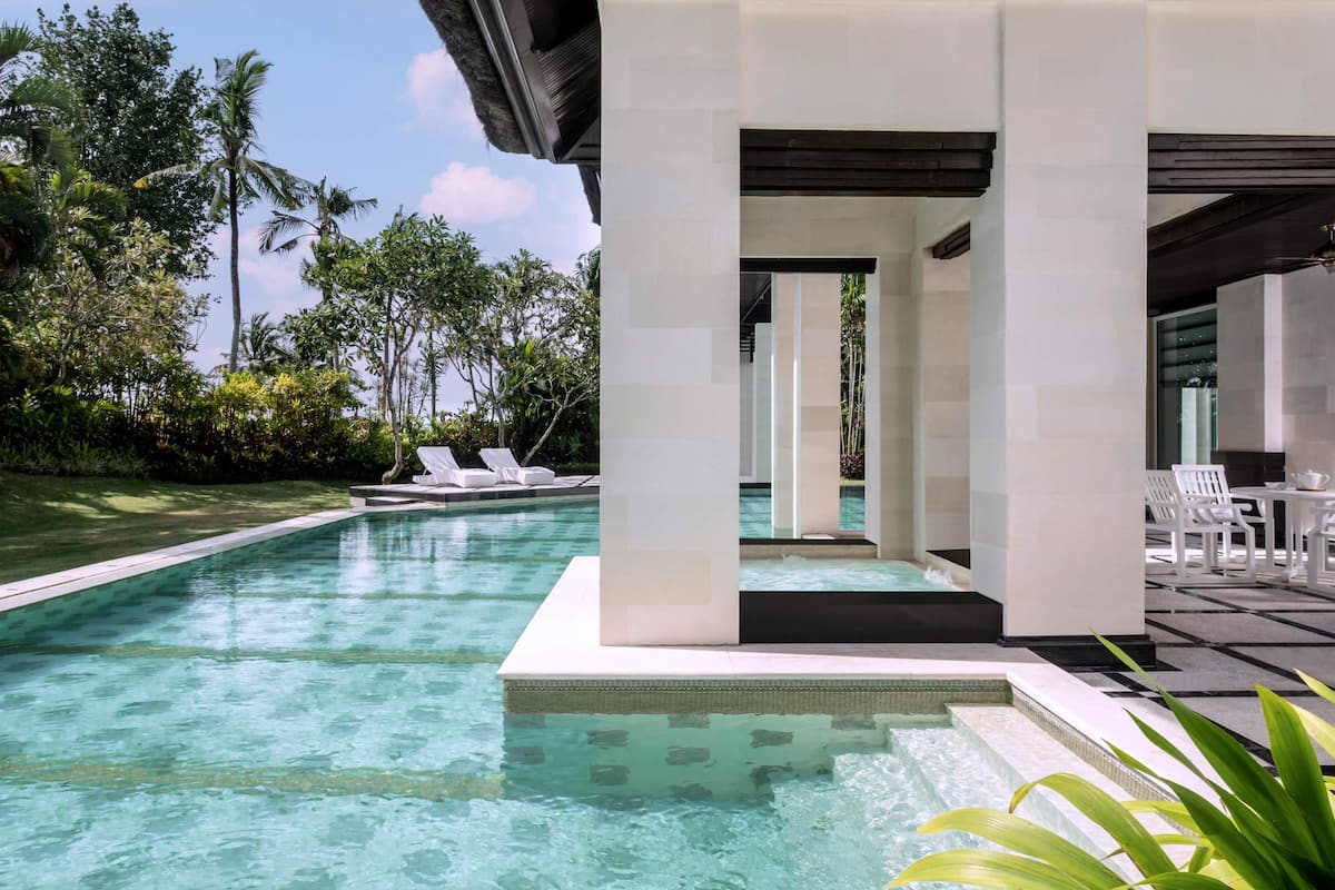 A swimming pool with clear water, surrounded by white columns and a covered seating area with white chairs.