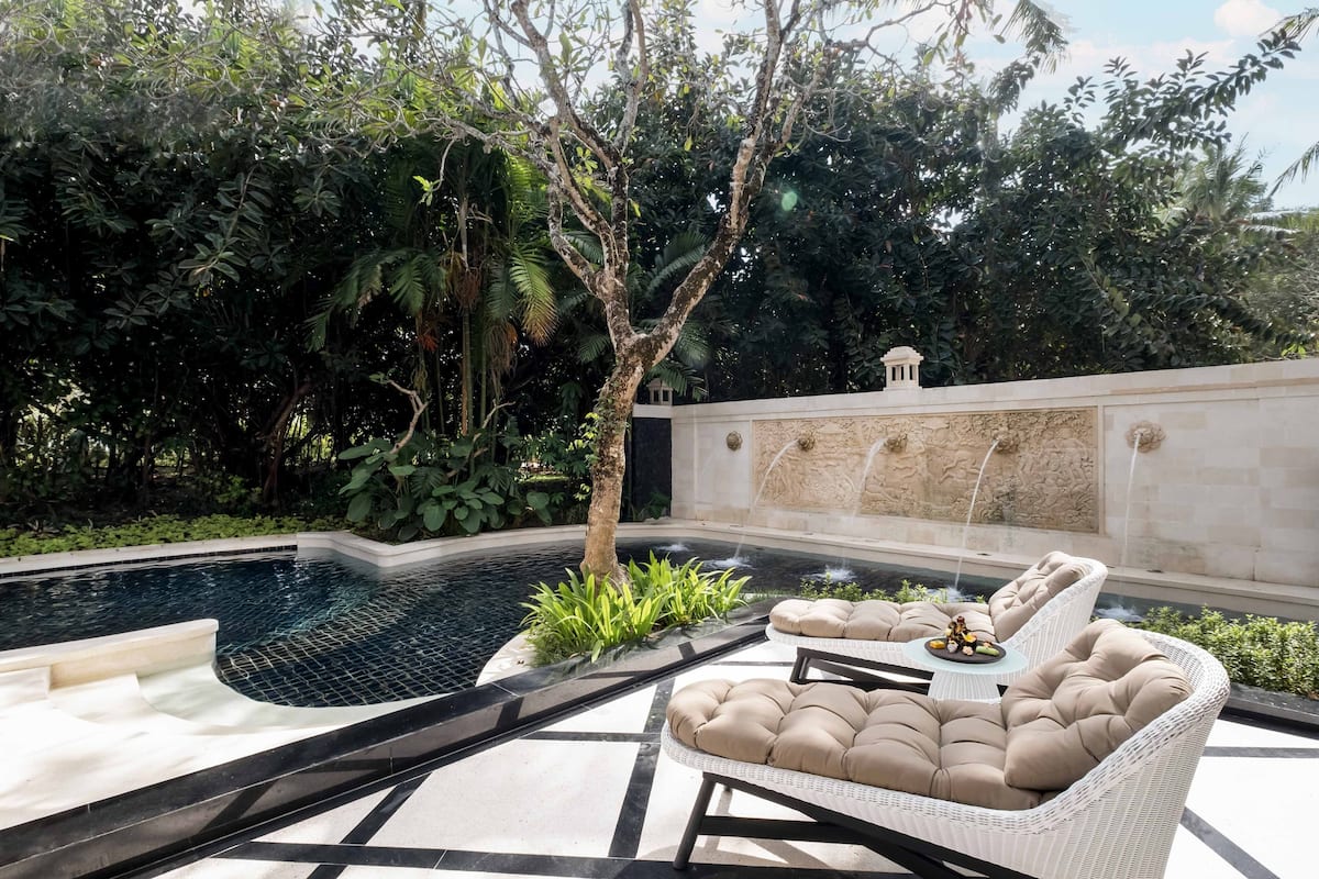 A pool area with a white wicker lounge chair, a small table with a decorative item, and a stone wall with a water feature.