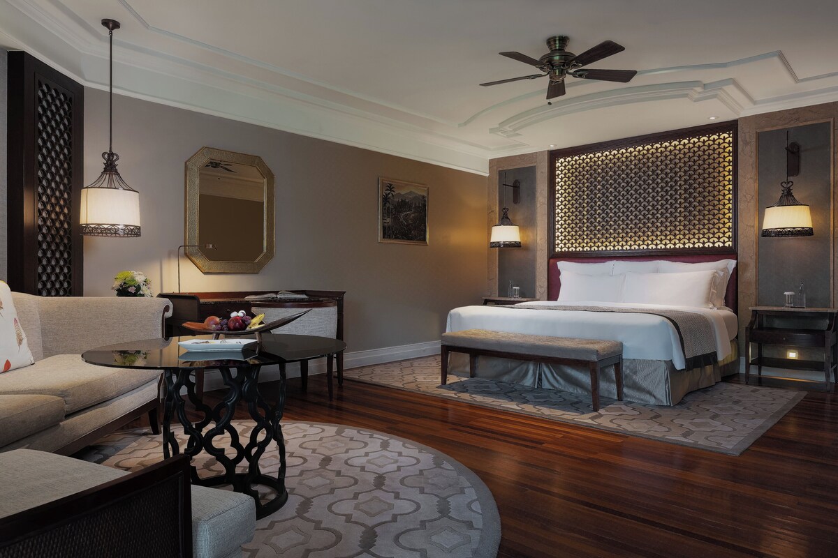 A hotel room with a bed, a sofa, a desk, a chair, a ceiling fan, and a patterned rug.