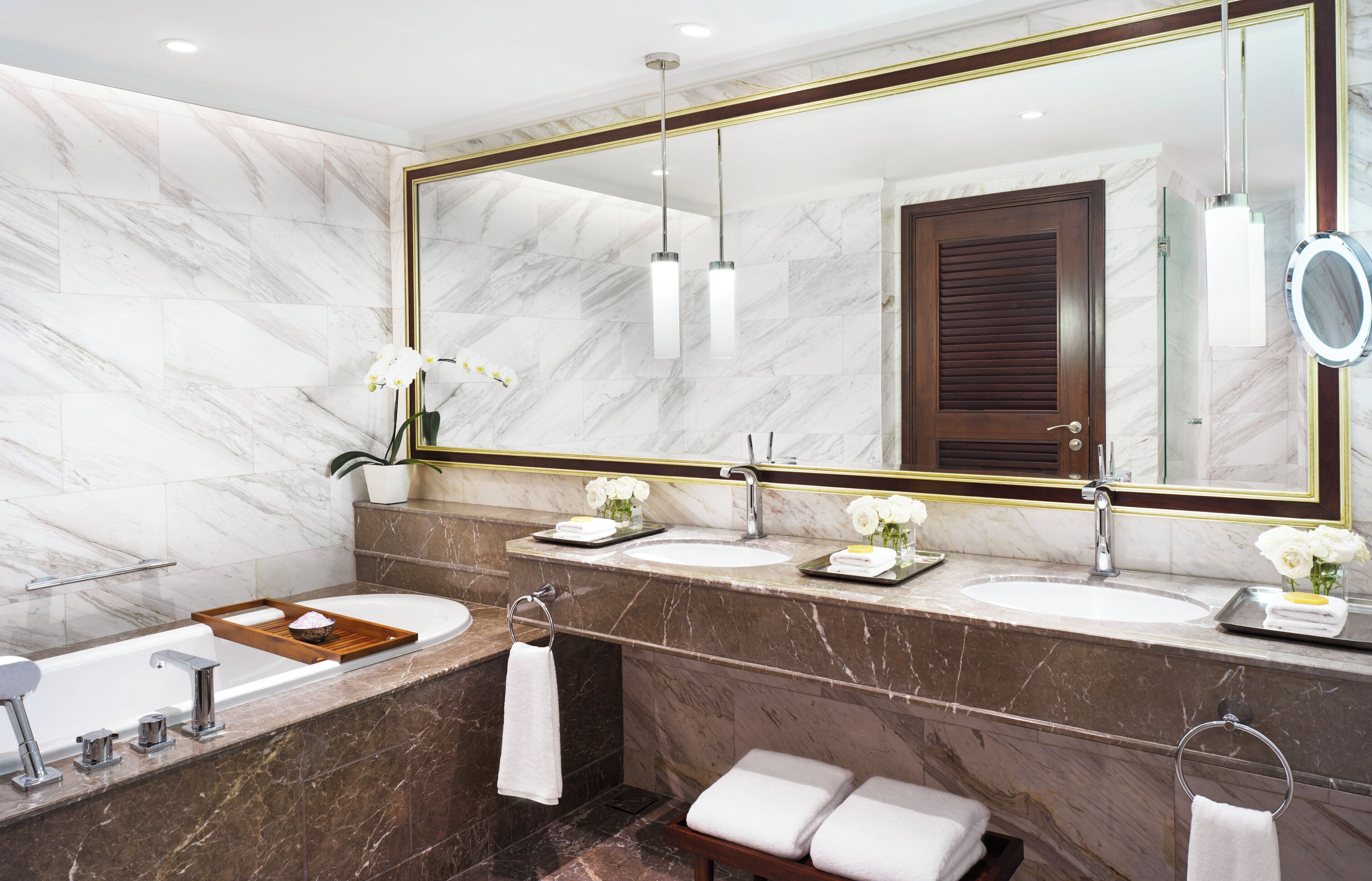 Une salle de bain moderne avec des murs en marbre, un grand miroir, deux lavabos, une baignoire et un banc.