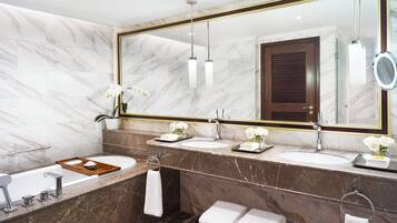 A modern bathroom with marble walls, a large mirror, two sinks, a bathtub, and a bench.