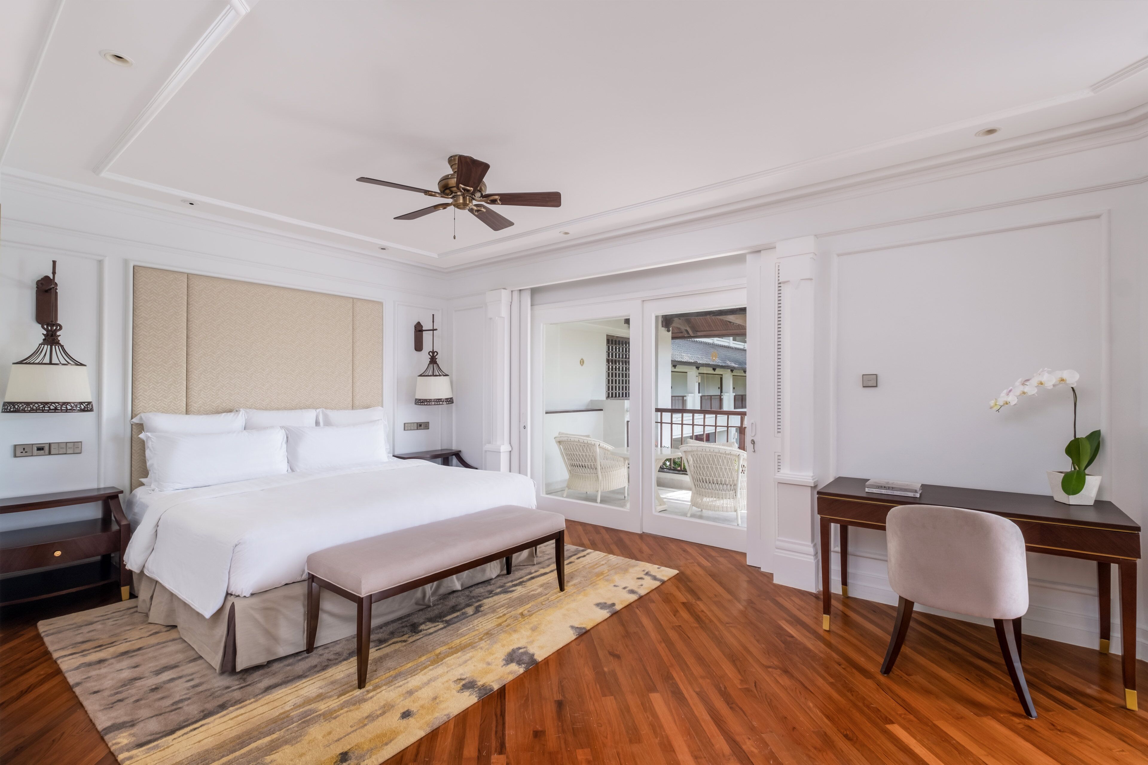 A hotel room with a large bed, a desk, a chair, a ceiling fan, and a balcony with wicker chairs.
