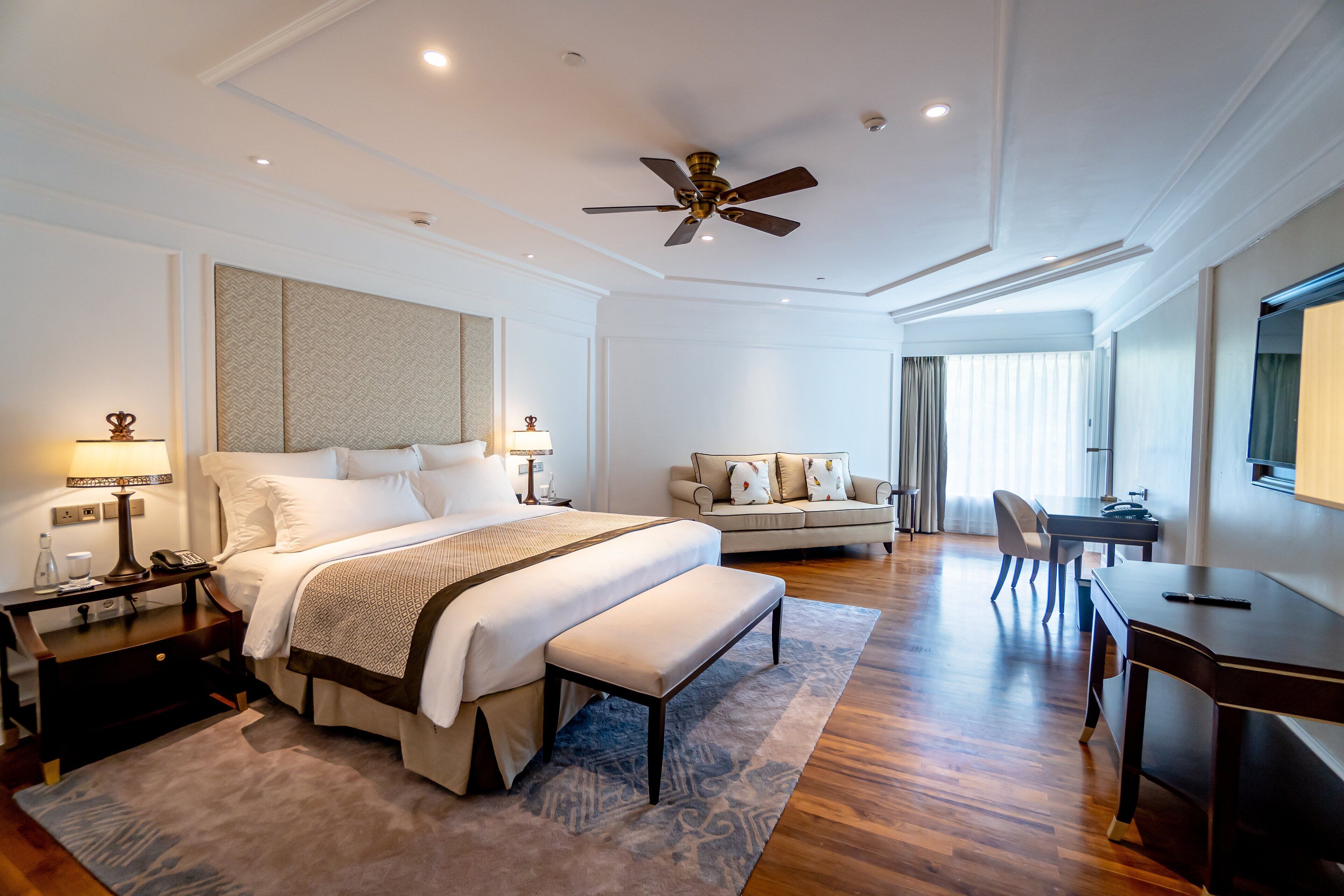 A modern hotel room with a large bed, a desk, a sofa, and a ceiling fan.